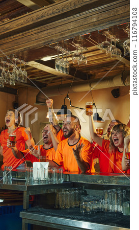 Friends in orange shirts cheer enthusiastically at bar, raising their glasses of beer, celebrating victory of favorite team. Friends in orange shirts cheer enthusiastically at bar, raising their glasses of beer, celebrating victory of favorite team. 116794108