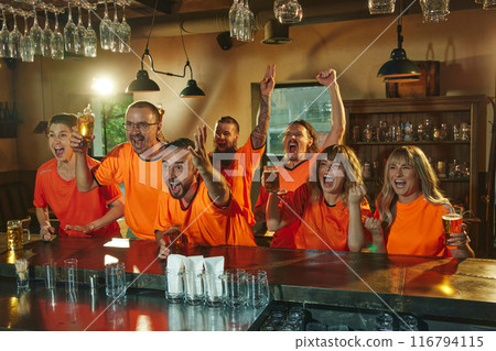 Enthusiastic fans in orange shirts sit at bar with glasses of beer, watching sports event and showing celebration expressions. Enthusiastic fans in orange shirts sit at bar with glasses of beer, watching sports event and showing celebration expressions. 116794115