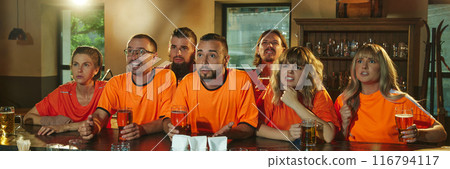 Banner. young people, women and men, in bright orange shirts sits and intensely watching TV and drinks beer at bar. Banner. young people, women and men, in bright orange shirts sits and intensely watching TV and drinks beer at bar. 116794117