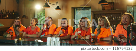 Banner. young people, women and men, in bright orange shirts sits and intensely watching TV and drinks beer at bar. Banner. young people, women and men, in bright orange shirts sits and intensely watching TV and drinks beer at bar. 116794122