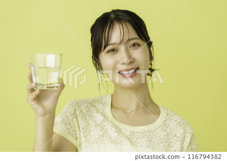 Portrait of a young woman holding a glass of water against a yellow background 116794382