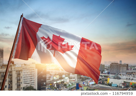 A red and white Canadian flag is flying high in the sky over a city 116794959
