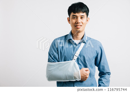 Confident businessman smiles despite broken arm, wearing supportive splint for treatment. Happy Asian man in sling, studio isolated on white, highlighting injury recovery. Copy space available. 116795744