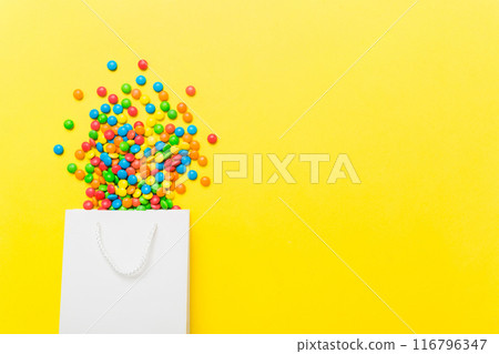 shopping paper gift bag in corner full of assorted traditional candies falling out on colored background with copy space. Happy Holidays sale concept 116796347