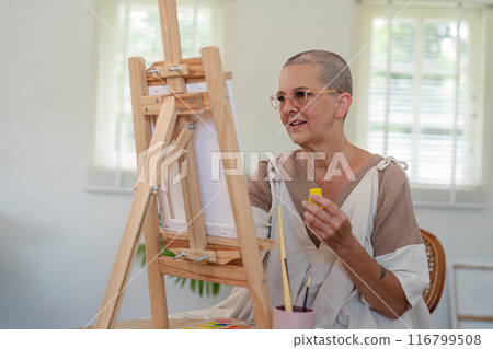 Elderly Woman Painting on Canvas in Bright Studio with Natural Light and Wooden Easel 116799508