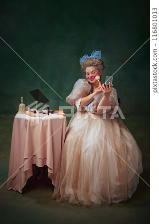 Portrait of young woman, princess sitting at table and making bright makeup against vintage green background. Full-length. 116801013