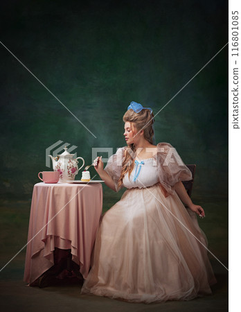Small bite. Portrait of elegant and, royal person in Rococo style dress sitting at table and making small bite of cake against vintage green background Small bite. Portrait of elegant and, royal person in Rococo style dress sitting at table and making small bite of cake against vintage green background 116801085