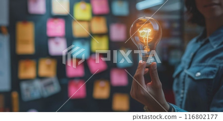 Businesswoman holding illuminated lightbulb with sticky notes in background 116802177