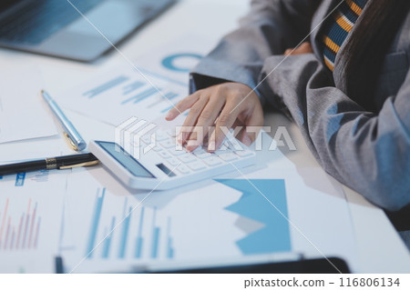 Pretty Asian woman sitting in the office, calculator, working on data, charts and documents on the table in business workspace. Looking camera. Pretty Asian woman sitting in the office, calculator, working on data, charts and documents on the table in business workspace. Looking camera. 116806134
