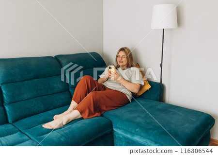 Young woman holding smartphone, using cellphone modern technology, checking cell phone apps, texting, watching video, browsing internet for shopping. Girl sitting on sofa at home. Full length body 116806544