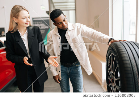Blonde saleswoman in business suit talking about characteristic of tires product to African-American customer young man came to look at assortment represented in auto service shop. 116808003