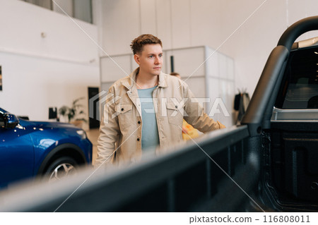 Young man buyer looking around big open trunk of car. Male customer choosing pickup truck in car dealership. Guy visitor walking in modern auto showroom and choosing new vehicle to buy or rent. 116808011