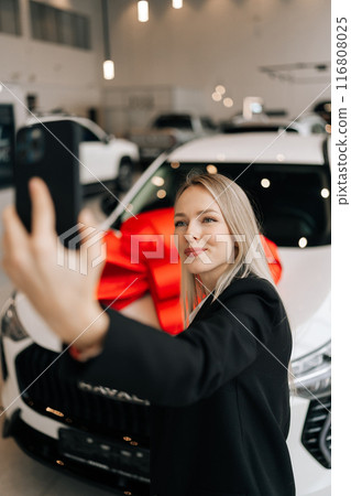 Vertical shot of happy female customer in black suit doing selfie shot pov on mobile phone choose auto want to buy new automobile in car showroom. Concept of choosing and buying new auto at showroom. Vertical shot of happy female customer in black suit doing selfie shot pov on mobile phone choose auto want to buy new automobile in car showroom. Concept of choosing and buying new auto at showroom. 116808025
