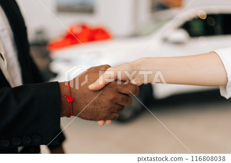 Closeup hands of unrecognizable black male dealer and female customer shaking hands in front of automobiles in car dealership center. Concept of choosing and buying new car at showroom office. Closeup hands of unrecognizable black male dealer and female customer shaking hands in front of automobiles in car dealership center. Concept of choosing and buying new car at showroom office. 116808038