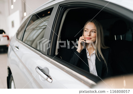 Satisfied successful businesswoman talking mobile phone, having friendly conversation. Cheerful blonde young woman sit in new car in automobile shop, renting buying auto luxury vehicle in dealership. 116808057