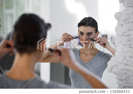 A young woman massages her face with gouache while looking in the mirror. 116808433