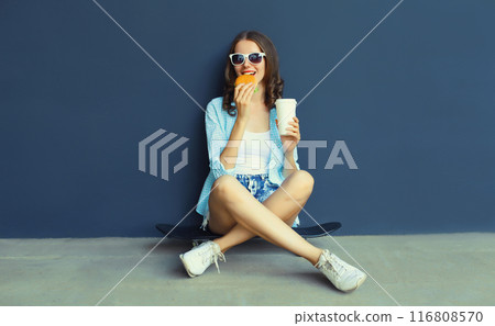 Happy modern young woman eating burger fast food, stylish teenage girl sitting on skateboard in city 116808570