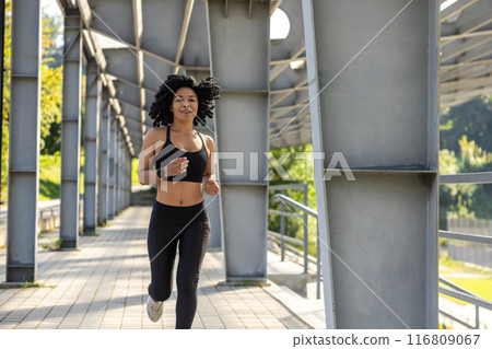 Curly-haired woman in black sportswear running and looking focused 116809067
