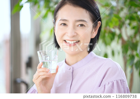 Smiling middle-aged woman drinking water in the living room Smiling middle-aged woman drinking water in the living room 116809738
