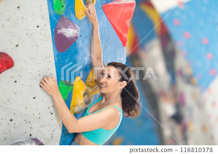 Adult woman is mastering climbing on training wall in gym, side view. Young woman holds on tightly to ledges and strives for top of bouldering route Adult woman is mastering climbing on training wall in gym, side view. Young woman holds on tightly to ledges and strives for top of bouldering route 116810378