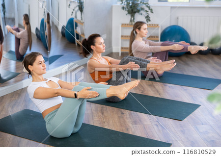 Diligent young women practicing upward boat pose of yoga on mat in fitness room Diligent young women practicing upward boat pose of yoga on mat in fitness room 116810529