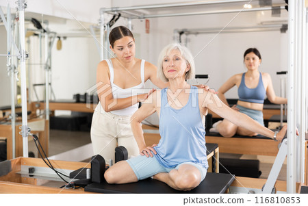 Female pilates instructor helping elderly woman doing exercises on reformer apparatus Female pilates instructor helping elderly woman doing exercises on reformer apparatus 116810853