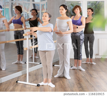 Elderly woman learning first ballet position at barre in group class Elderly woman learning first ballet position at barre in group class 116810915