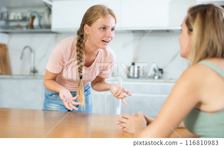 Smiling teenage girl talking friendly with woman in home kitchen Smiling teenage girl talking friendly with woman in home kitchen 116810983