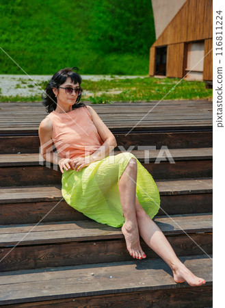 50 years old russian woman in short dress and sunglasses sitting on a stairs and enjoying on the summer day  116811224