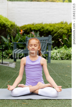 Vertical of happy biracial girl sitting in garden, practicing yoga meditation, with copy space 116811313