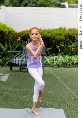 Vertical of happy biracial girl standing on one leg in garden, practicing yoga, with copy space 116811330