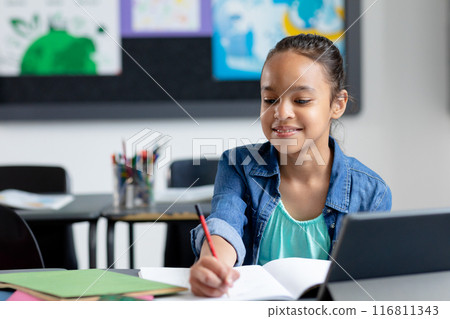 Smiling biracial schoolgirl sitting at desk with tablet, writing in class, with copy space Smiling biracial schoolgirl sitting at desk with tablet, writing in class, with copy space 116811343