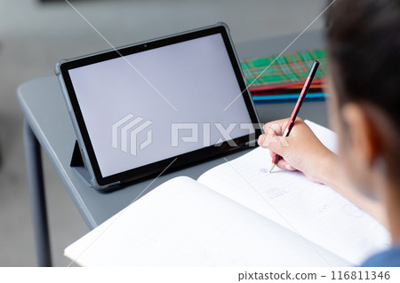 Biracial schoolgirl sitting at desk writing in class and using tablet, with copy space on screen 116811346