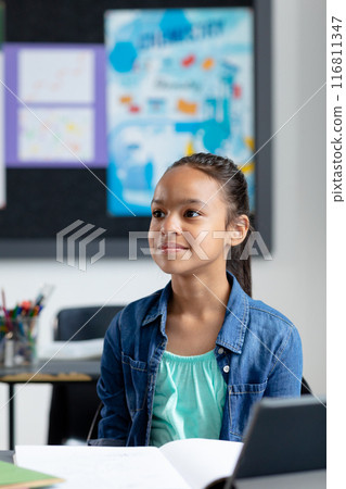 Vertical of happy biracial schoolgirl sitting at desk in class, listening, with copy space Vertical of happy biracial schoolgirl sitting at desk in class, listening, with copy space 116811347