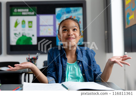 Biracial schoolgirl shrugs, smiles at desk with copy space 116811361