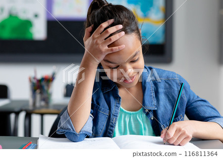 Happy biracial schoolgirl sitting at desk in class, resting head on hand and writing, copy space 116811364