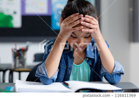 Stressed biracial schoolgirl sitting at desk in class, head in hands, staring at book, copy space 116811369