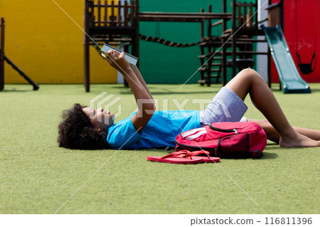 Smiling biracial schoolgirl lying on her back in school playground reading book, with copy space 116811396