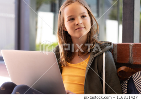 Smiling caucasian schoolgirl using laptop, sitting outside school, with copy space 116811427
