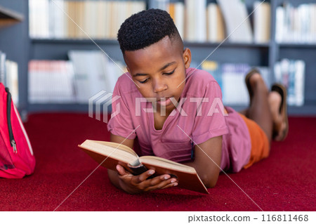 African american schoolboy lying on floor reading book in school library, concentrating, copy space African american schoolboy lying on floor reading book in school library, concentrating, copy space 116811468