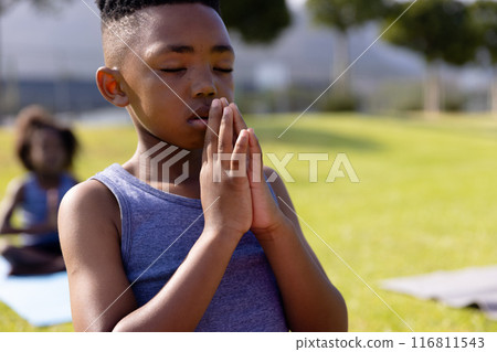 Happy african american schoolboy doing yoga and meditating on field at school 116811543