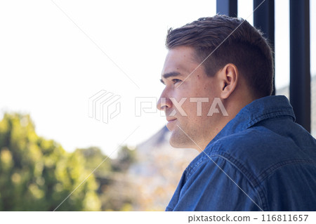 Young man in denim shirt gazing thoughtfully out window, enjoying nature view, copy space Young man in denim shirt gazing thoughtfully out window, enjoying nature view, copy space 116811657