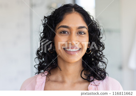 Smiling woman with curly hair wearing pink shirt, looking at camera indoors 116811662