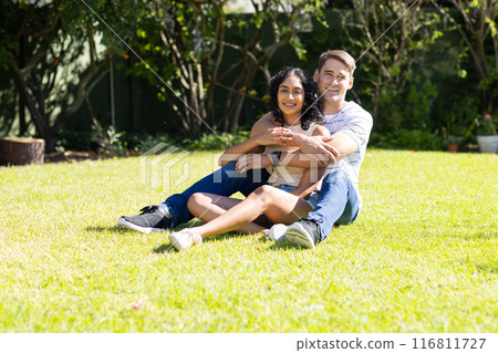 Sitting on grass, young couple hugging and smiling in sunny garden Sitting on grass, young couple hugging and smiling in sunny garden 116811727