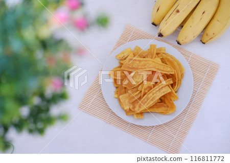 Crispy banana chips on a white plate with fresh bananas. Crispy banana chips on a white plate with fresh bananas. 116811772