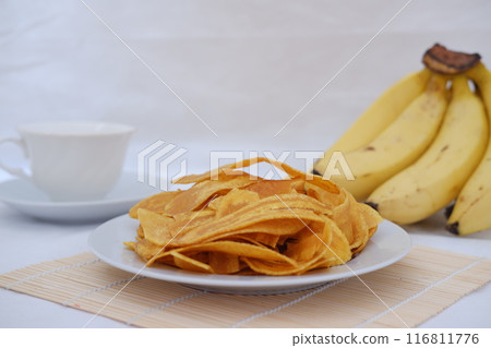 Crispy banana chips on a white plate with fresh bananas. Crispy banana chips on a white plate with fresh bananas. 116811776