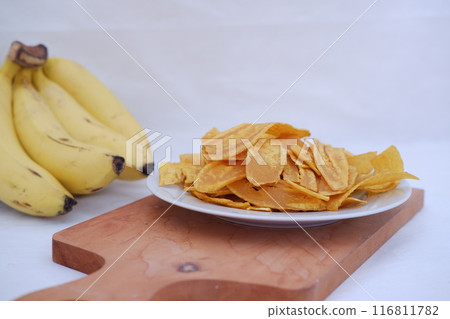 Crispy banana chips on a white plate with fresh bananas. Crispy banana chips on a white plate with fresh bananas. 116811782