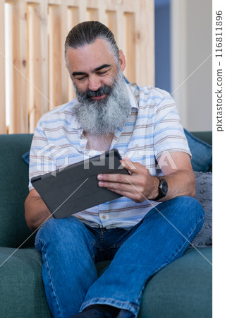 Using tablet, senior man with gray beard sitting on couch, smiling and reading Using tablet, senior man with gray beard sitting on couch, smiling and reading 116811896