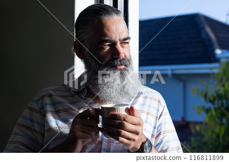 Holding cup, senior man with beard enjoying morning sunlight indoors Holding cup, senior man with beard enjoying morning sunlight indoors 116811899