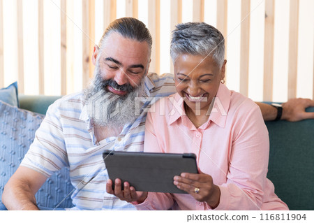 Senior couple sitting on couch, smiling and using tablet together Senior couple sitting on couch, smiling and using tablet together 116811904